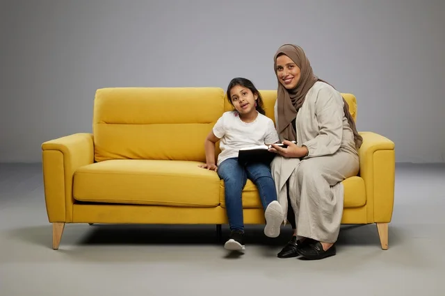 أم سعودية تقرأ كتابًا مع ابنتها على أريكة صفراء