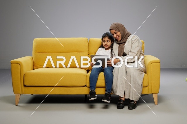 أم وابنتها تستخدمان جهازاً لوحياً على أريكة صفراء