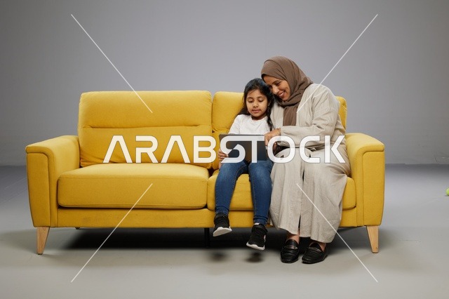 أم وابنتها تستخدمان جهازاً لوحياً على أريكة صفراء