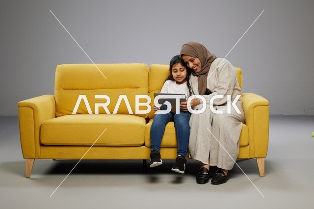 أم وابنتها تستخدمان جهازاً لوحياً على أريكة صفراء