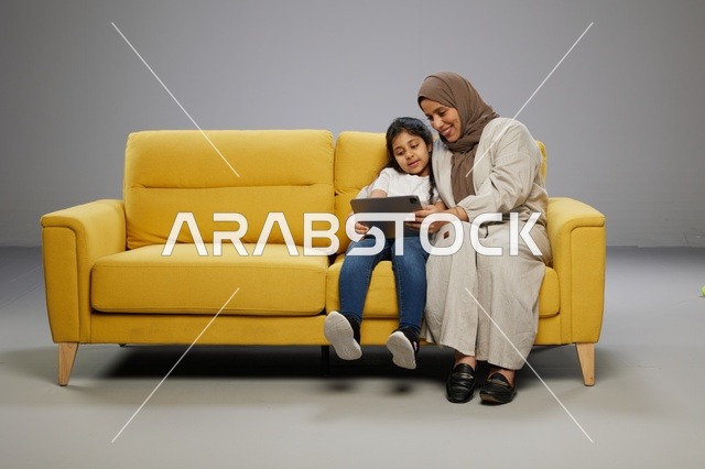 أم وابنتها تستخدمان جهازاً لوحياً على أريكة صفراء