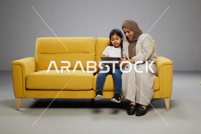 أم وابنتها تستخدمان جهازاً لوحياً على أريكة صفراء