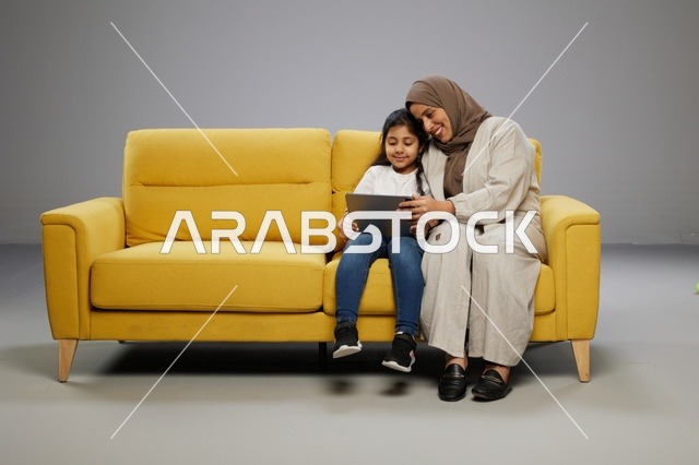 أم وابنتها تستخدمان جهازاً لوحياً على أريكة صفراء