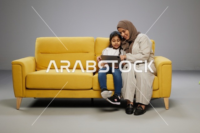 أم وابنتها تستخدمان جهازاً لوحياً على أريكة صفراء