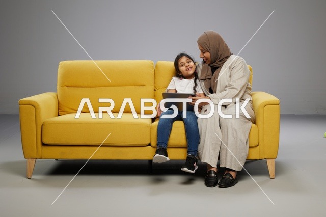 أم وابنتها تستخدمان جهازًا لوحيًا على أريكة صفراء