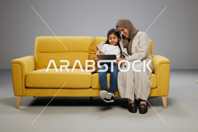 أم عربية وابنتها تستخدمان جهاز لوحي على أريكة صفراء