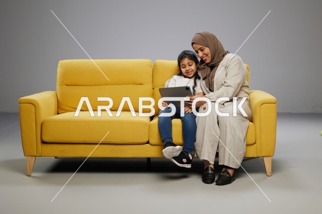 أم وابنتها تستخدمان جهازاً لوحياً على أريكة صفراء