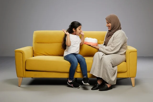 Arab Mother Surprising Daughter with Cake on Yellow Sofa