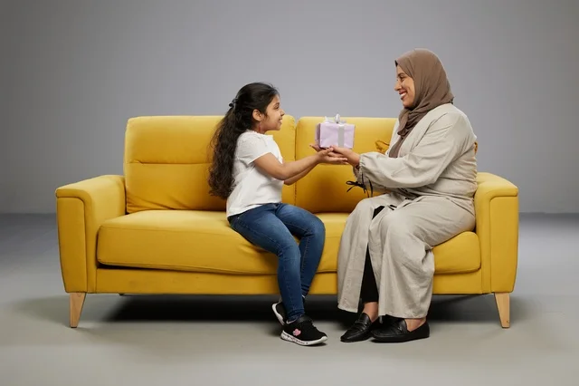 Arab Mother Giving Gift to Daughter on Yellow Sofa