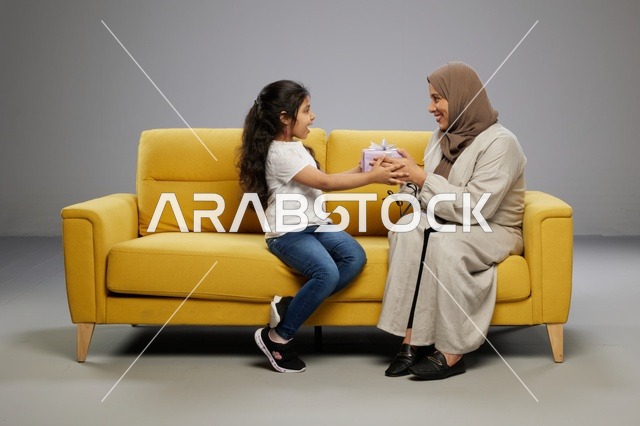 أم عربية تقدم هدية لابنتها الصغيرة على أريكة صفراء في استوديو