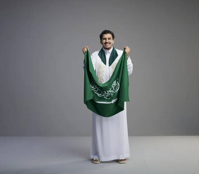 شاب سعودي مبتسم يحمل العلم الوطني السعودي في استوديو