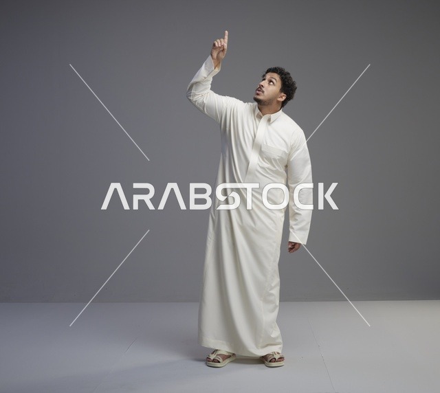رجل سعودي بالثوب الأبيض يشير للأعلى بخلفية رمادية