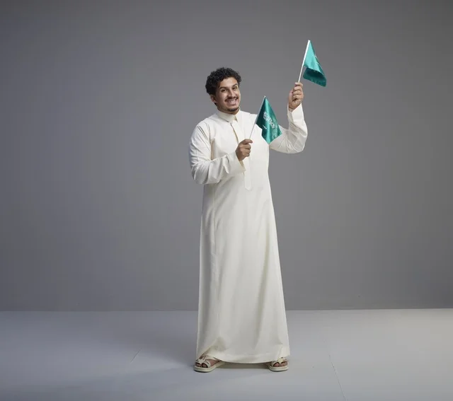 شاب سعودي مبتسم يحمل أعلام المملكة في الاستوديو