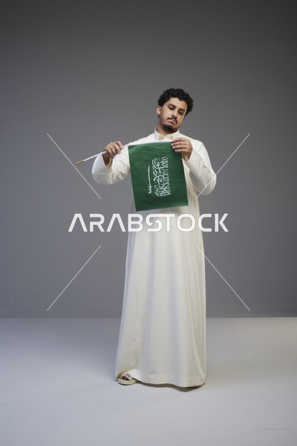 شاب سعودي بالثوب التقليدي يمسك علم السعودية في استوديو