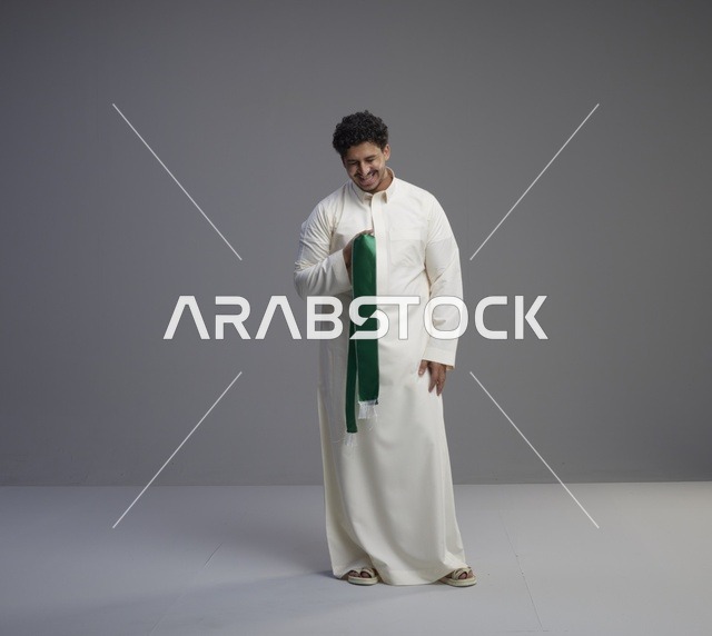 رجل سعودي بالثوب يحمل وشاحاً وطنياً أخضر في استوديو