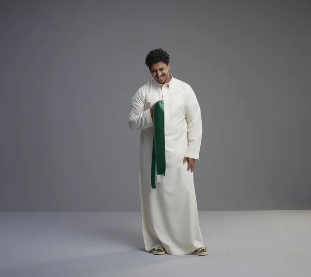 رجل سعودي بالثوب يحمل وشاحاً وطنياً أخضر في استوديو