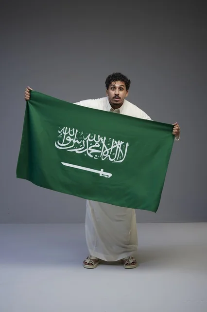 رجل سعودي يحمل علم المملكة العربية السعودية بخلفية رمادية
