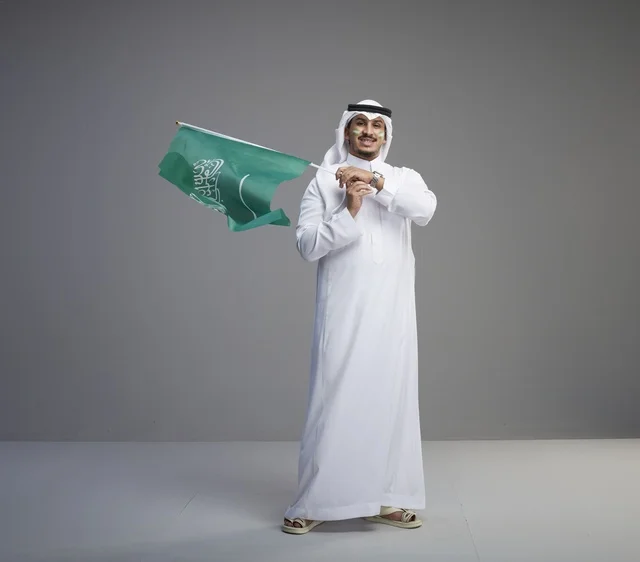 رجل سعودي يحمل العلم الوطني في خلفية استوديو رمادية
