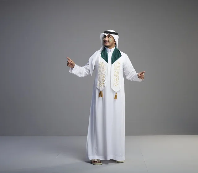 خريج سعودي بالزي التقليدي يشير للأعلى في استوديو رمادي