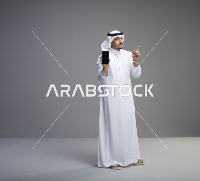 رجل سعودي يحمل هاتفاً ذكياً بشاشة فارغة بالثوب التقليدي