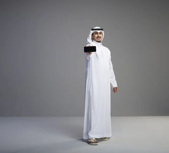 رجل سعودي يحمل هاتف ذكي بشاشة فارغة في استوديو رمادي