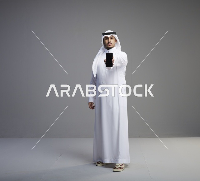 رجل سعودي يحمل هاتف ذكي بشاشة فارغة بخلفية رمادية