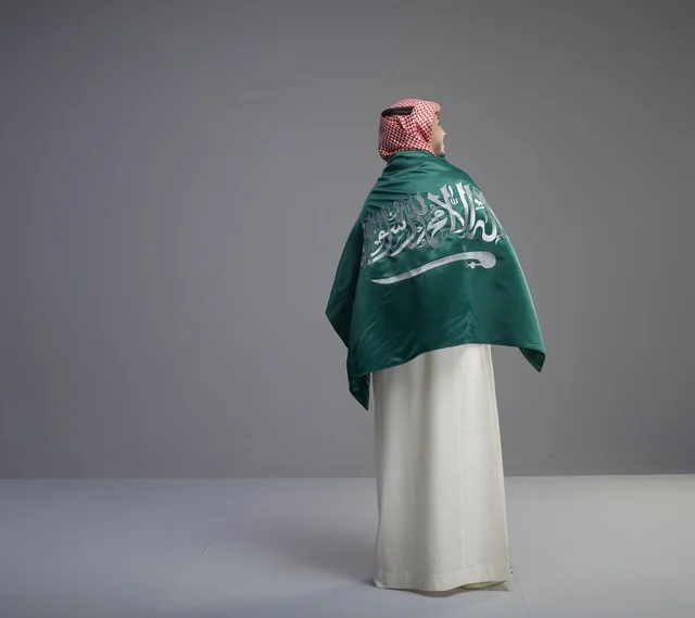 رجل سعودي يلتف بعلم المملكة العربية السعودية بخلفية رمادية