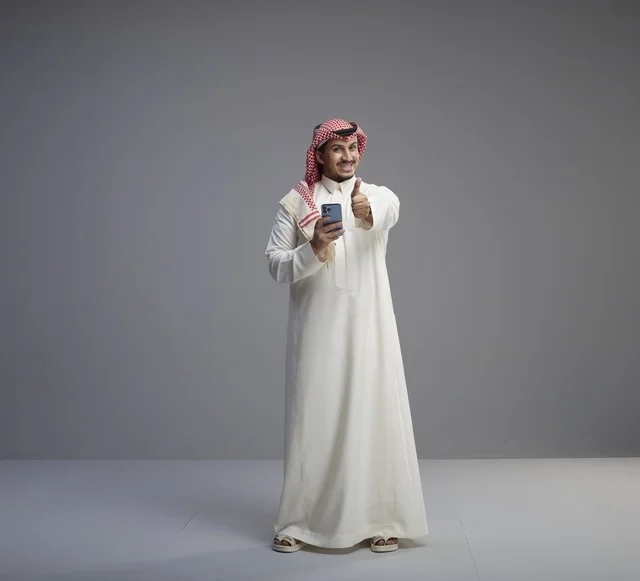 رجل سعودي مبتسم يستخدم الهاتف الذكي مع إيماءة الإبهام