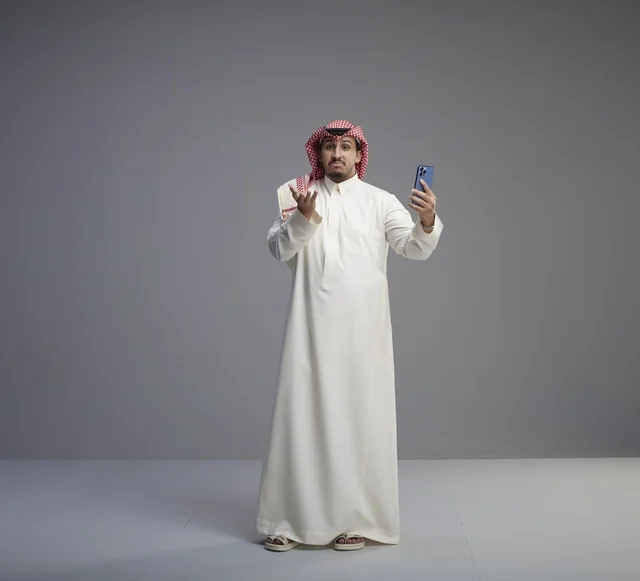 رجل سعودي يستخدم هاتفاً ذكياً بتعبير وجه حائر في استوديو