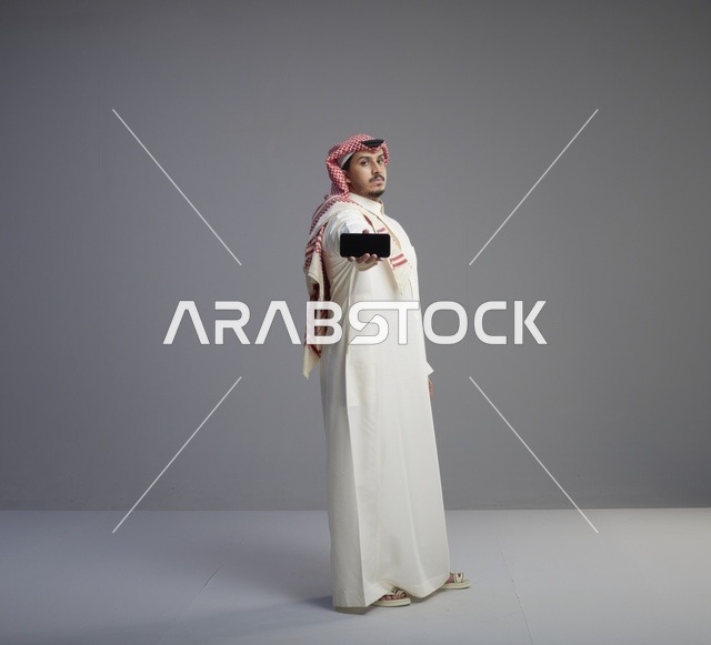 رجل سعودي يرتدي الثوب يحمل هاتف ذكي بخلفية رمادية