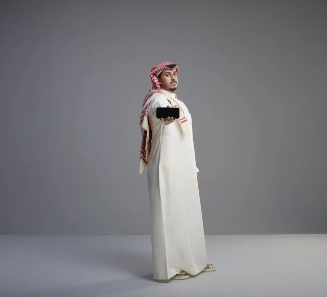رجل سعودي يرتدي الثوب يحمل هاتف ذكي بخلفية رمادية