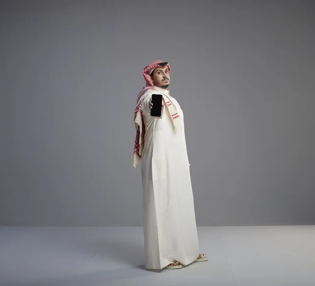 رجل سعودي يحمل هاتفاً ذكياً في استوديو بخلفية رمادية