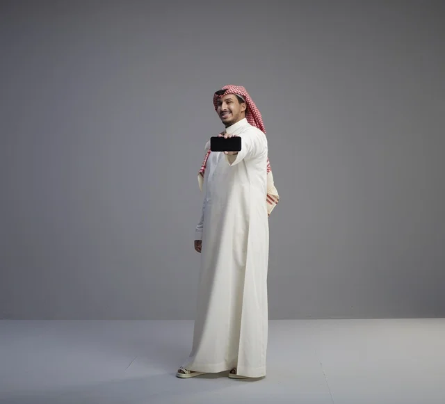 رجل سعودي يحمل نموذج هاتف ذكي بخلفية رمادية