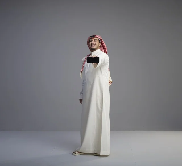 رجل سعودي يحمل نموذج هاتف ذكي أمام خلفية رمادية