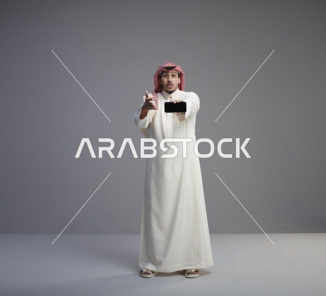 رجل سعودي يحمل هاتف ذكي بتعبيرات وجه مندهشة في الاستوديو