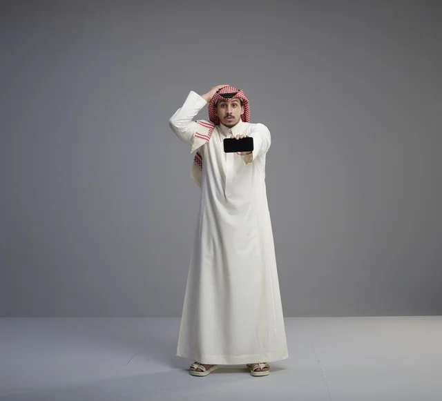 رجل سعودي يحمل هاتف ذكي بتعبيرات وجه مندهشة
