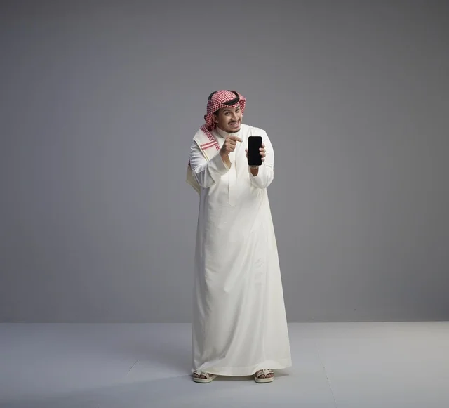 رجل سعودي يحمل نموذج هاتف ذكي في استوديو بخلفية رمادية