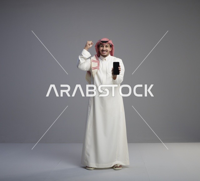 رجل سعودي يحمل هاتف ذكي مع إيماءة فوز في استوديو