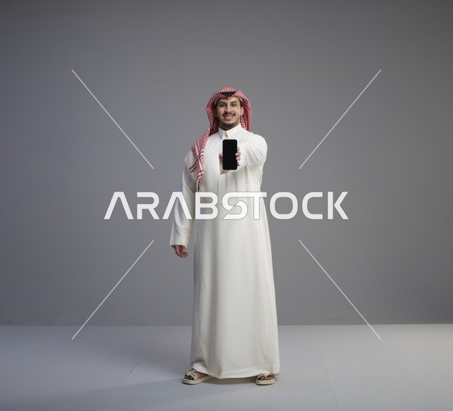 رجل سعودي يحمل هاتف ذكي في استوديو بخلفية رمادية