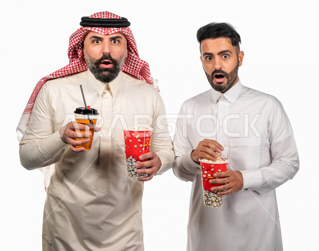 Portrait of two Saudi Arabian Gulf men carrying a box of popcorn, gestures of the face and hands indicate enthusiasm and amazement, watching movies inside the cinema, watching horror and action movies, movies and cinema