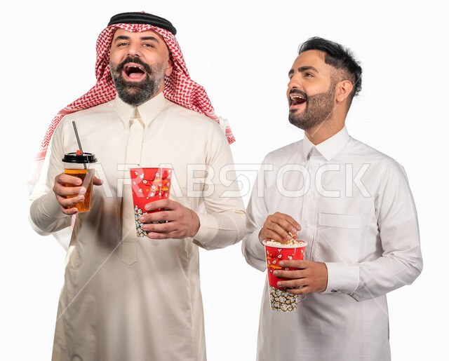 Portrait of two Saudi Arabian Gulf men carrying a box of popcorn, gestures of the face and hands indicate fun and enjoyment, watching movies inside the cinema, watching movies and comic series, movies and cinema