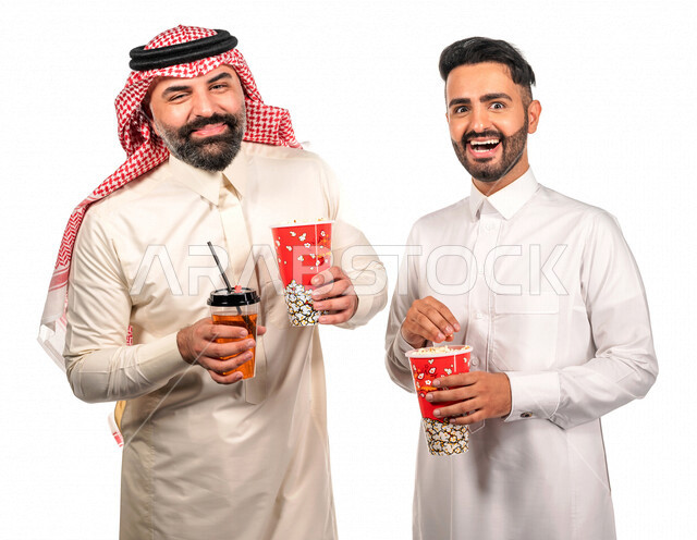 Portrait of two Saudi Arabian Gulf men carrying a box of popcorn, gestures of the face and hands indicate fun and enjoyment, watching movies inside the cinema, watching movies and comic series, movies and cinema