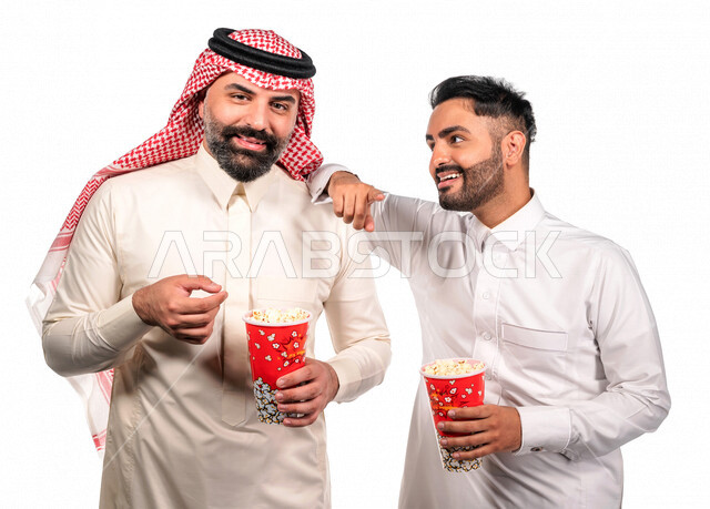 Portrait of two Saudi Arabian Gulf men carrying a box of popcorn, gestures of the face and hands indicate fun and enjoyment, watching movies inside the cinema, watching movies and comic series, movies and cinema