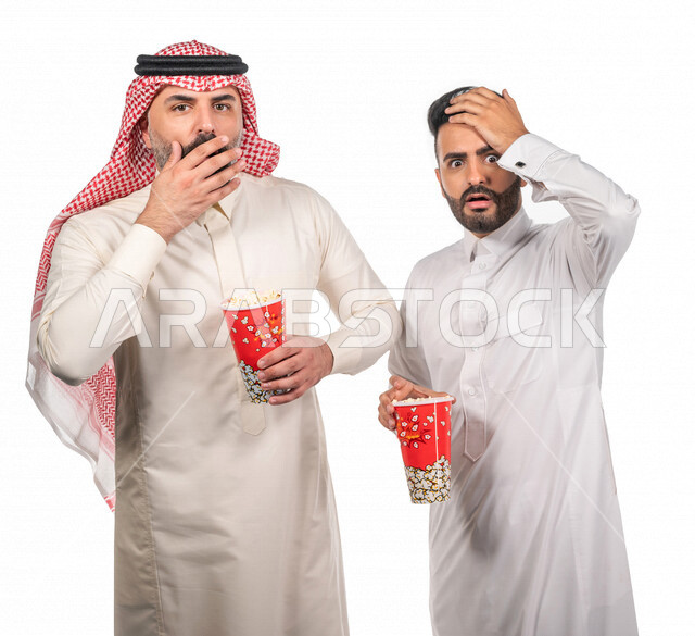 Portrait of two Saudi Arabian Gulf men carrying a box of popcorn, gestures of the face and hands indicate enthusiasm and amazement, watching movies inside the cinema, watching horror and action movies, movies and cinema