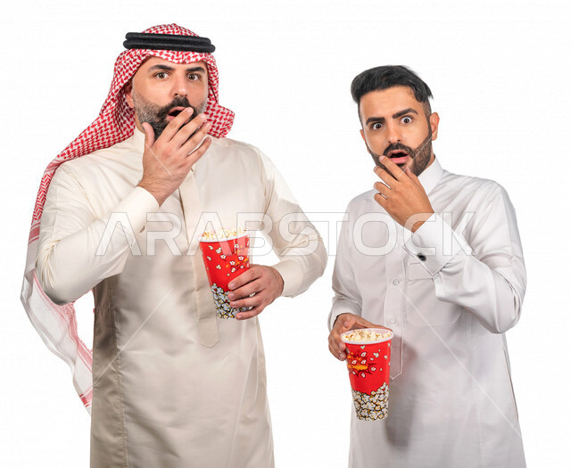 Portrait of two Saudi Arabian Gulf men carrying a box of popcorn, gestures of the face and hands indicate enthusiasm and amazement, watching movies inside the cinema, watching horror and action movies, movies and cinema