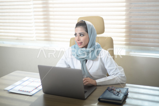 A Saudi Arabian Gulf doctor wearing a medical coat and sitting in front of a laptop, providing online medical consultations, following up on the patient’s medical record, a satisfactory case study, the concept of medicine and health care