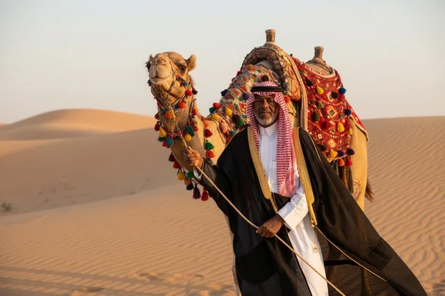 رجل سعودي مع جمل مزين في الصحراء وقت الغروب