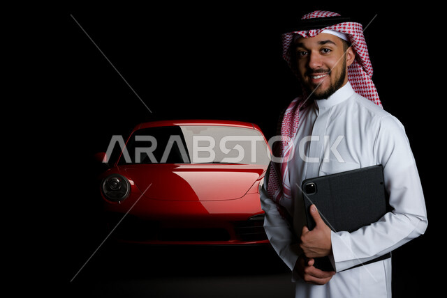 بورتريه لرجل عربي خليجي سعودي مبتسم يحمل بيديه جهاز تقني ذكي ( تابلت ) يقف بجانب سيارة حمراء ، مدير مبيعات بشركة سيارات ، تقديم استشارات و توصيات للعملاء ، جمع بيانات لاتجاهات السوق ، منتجات تنافسية ، رفاهية و غنى