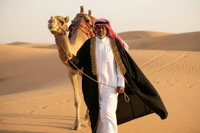 رجل سعودي بالبشت التقليدي يقود جملاً في الصحراء