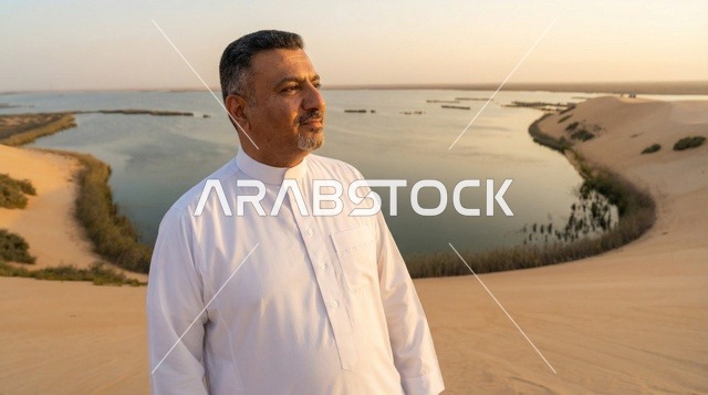 رجل سعودي بالثوب التقليدي عند بحيرة صحراوية وقت الغروب
